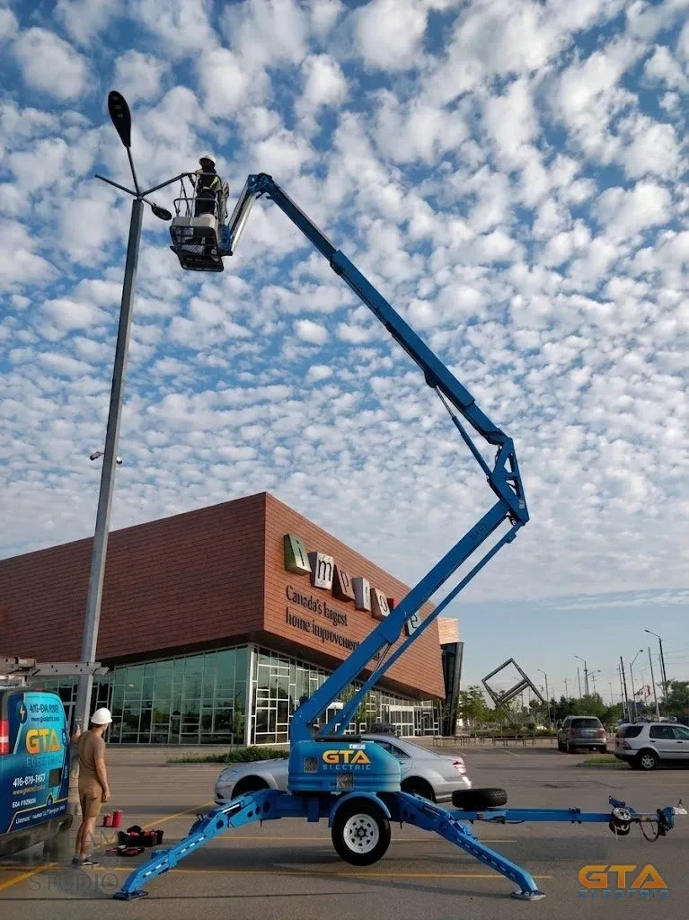 GTA Electric electrician working from an aerial boom lift to service a parking-lot light standard outside Improve Canada in Vaughan, with the branded GTA Electric van and lift on site